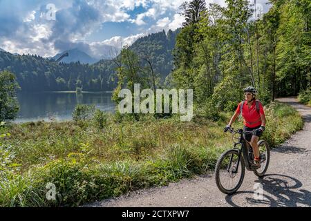 Nette ältere Frau mit ihrem Elektro-Mountainbike über dem Freiberger See und einer großen Skiflugschanze in den Allgauer Alpen bei Oberstdorf, Bayern, Deutschland Stockfoto