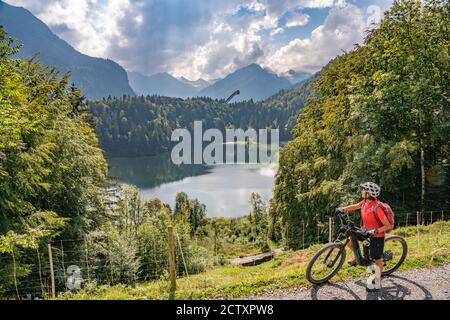 Nette ältere Frau mit ihrem Elektro-Mountainbike über dem Freiberger See und einer großen Skiflugschanze in den Allgauer Alpen bei Oberstdorf, Bayern, Deutschland Stockfoto