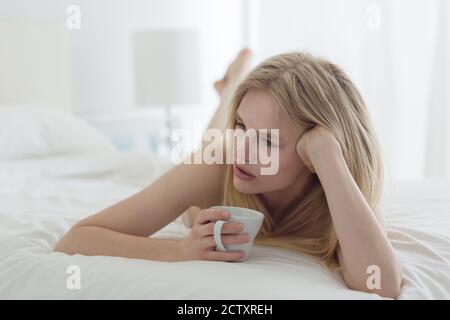 Nachdenkliche junge blonde Frau, die Kaffee trinkt, während sie im Bett liegt. Stockfoto