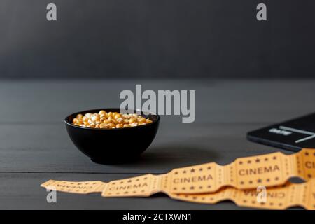 Schüssel mit Maiskörnern, zum Zubereiten von Popcornen, neben einem Klappbrett auf schwarzem Holzhintergrund. Film- und Fernsehindustrie, selektiver Fokus, conce
