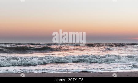 Sonnenuntergang Himmel mit stürmischen weißen glänzenden Wellen am Meer Sandstrand in Pastelltönen Stockfoto