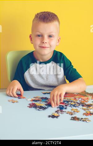 Portrait eines niedlichen Jungen, der am Tisch sitzt und Puzzles zusammenstellt. Frühzeitiges Lernen. Auf gelbem Hintergrund Stockfoto