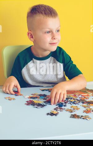 Portrait eines niedlichen Jungen, der am Tisch sitzt und Puzzles zusammenstellt. Frühzeitiges Lernen. Auf gelbem Hintergrund Stockfoto