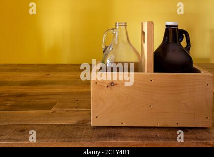 Zwei leere Glas-halbe Gallonen-Krüge, ein Bernstein, einer klar, in einer rustikalen Holzkiste auf einem Holztisch mit einem hellgelben Hintergrund mit Kopierraum Stockfoto