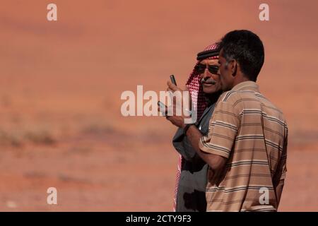 Wadi Rum, Jordanien 04/01/2010: Zwei Männer unterhalten sich mitten in der Wüste. Sie zeigen sich Handys zueinander. Einer trägt trad Stockfoto
