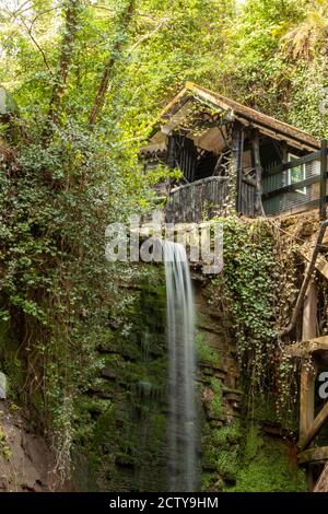 Langzeitbelichtung eines kleinen Wasserfalls. Es gibt ein Holzhaus auf der Spitze der Klippe am Rande der Felswand. Die Steinmauer hinter dem Fall ist Stockfoto