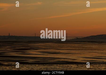 Low-Light-Aufnahme bei Sonnenuntergang am Ufer des Flusses Tejo, der zwischen Almada und Lissabon liegt. Es zeigt Silhouetten beider Städte, die 25 de abr Stockfoto