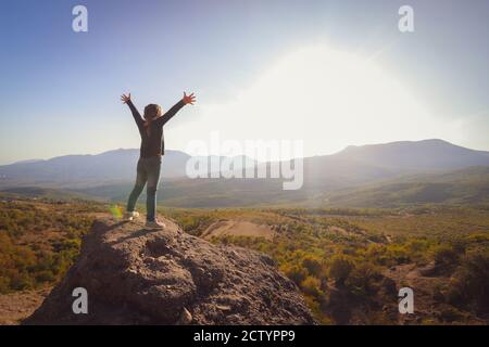 Kleines Mädchen auf einem Felsen in den Bergen. Das Kind streckte seine Arme zur Sonne. Hochwertige Fotos Stockfoto
