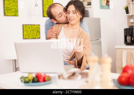 Ehemann küsst Frau Wange beim Lesen Ideen für Frühstück in der Küche. Glücklich liebend fröhlich romantisch in Liebe Paar zu Hause mit modernen WiFi-WLAN-Internet-Technologie Stockfoto