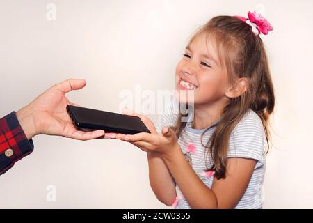 Vater schenkt seiner Tochter ein Geschenk an ihrem Geburtstag. Die Hand eines Mannes übergibt dem Mädchen in Nahaufnahme ein schwarzes neues Smartphone. Ein glückliches Kind packt das Telefon Stockfoto