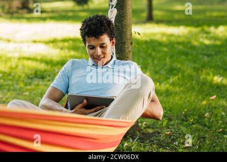 Junger Mann liegt in einer Hängematte im Park und Mit einem digitalen Tablet Stockfoto