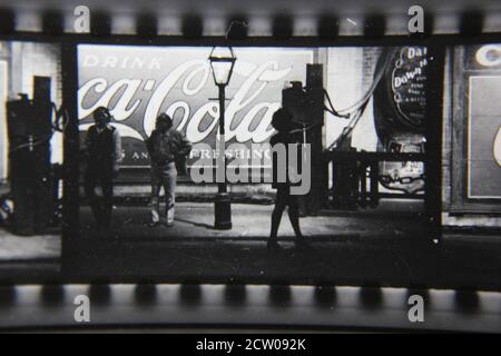 Feine Vintage-Schwarz-Weiß-Fotografie der 1970er Jahre von Profilen von Stadtmenschen, die vor einer riesigen Coca-Cola-Anzeige auf einer Wand gemalt gehen. Stockfoto