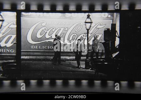Feine Vintage-Schwarz-Weiß-Fotografie der 1970er Jahre von Profilen von Stadtmenschen, die vor einer riesigen Coca-Cola-Anzeige auf einer Wand gemalt gehen. Stockfoto