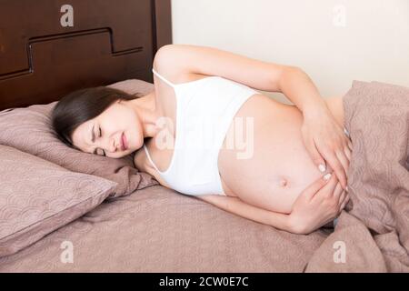 Gestresste schwangere Frau im Bett liegend mit der Hand auf dem Bauch, Grimacing von der Schraube. Stockfoto