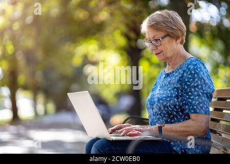 Ältere Frau mit Laptop auf der Bank im Freien Stockfoto