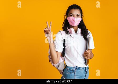 Junge glückliche Mädchen Student trägt eine medizinische Schutzmaske, hält Laptop-Computer isoliert auf einem gelben Hintergrund Stockfoto