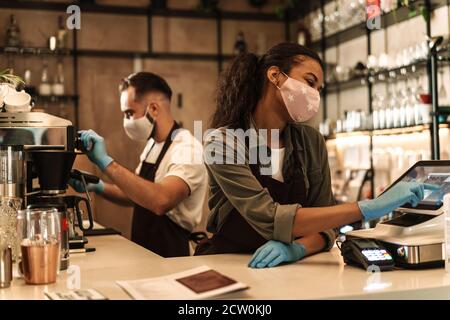 Zwei Baristas mit medizinischer Maske, die Kaffee hinter der Theke servieren Während der Quarantäne in Innenräumen Stockfoto