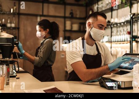 Zwei Baristas mit medizinischer Maske, die Kaffee hinter der Theke servieren Während der Quarantäne in Innenräumen Stockfoto