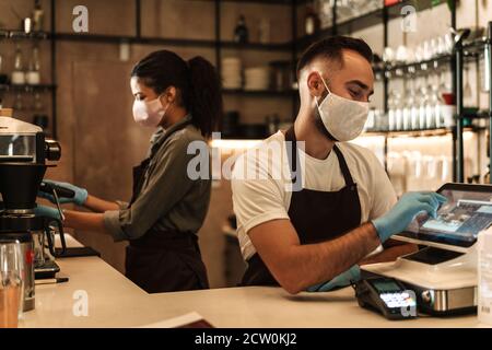 Zwei Baristas mit medizinischer Maske, die Kaffee hinter der Theke servieren Während der Quarantäne in Innenräumen Stockfoto
