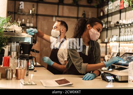 Zwei Baristas mit medizinischer Maske, die Kaffee hinter der Theke servieren Während der Quarantäne in Innenräumen Stockfoto