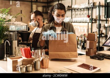 Zwei Baristas mit medizinischer Maske, die Kaffee hinter der Theke servieren Während der Quarantäne in Innenräumen Stockfoto