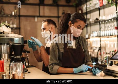 Zwei Baristas mit medizinischer Maske, die Kaffee hinter der Theke servieren Während der Quarantäne in Innenräumen Stockfoto