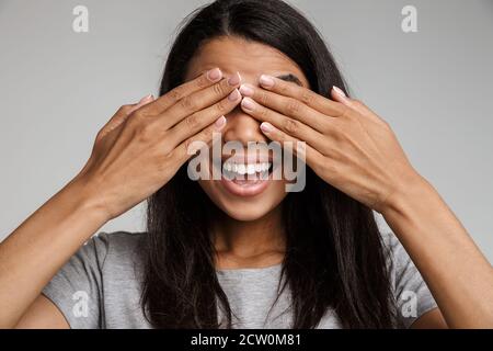 Aufgeregt junge afro-amerikanische Frau trägt Freizeitkleidung isoliert auf grauem Hintergrund stehend, bedeckt Gesicht mit Händen Stockfoto