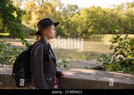Junge kaukasische Frau in einer Lederjacke und Hut steht Am Teich Stockfoto