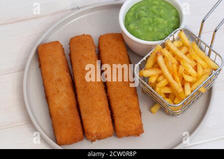 Fischfinger mit Chips in einem Drahtkorb und matschigen Erbsen serviert. Weißer Holzhintergrund Stockfoto
