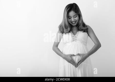 Studio Aufnahme von jungen glücklich asiatische schwangere Frau lächeln während Auf ihren Bauch schauen und Herz Hand Zeichen Stockfoto
