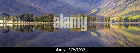 Die berühmte Char Hütte und die Wachtbäume in Buttermere spiegeln sich in den ruhigen Gewässern, während das Morgenlicht die umliegenden Fjells beleuchtet. Stockfoto