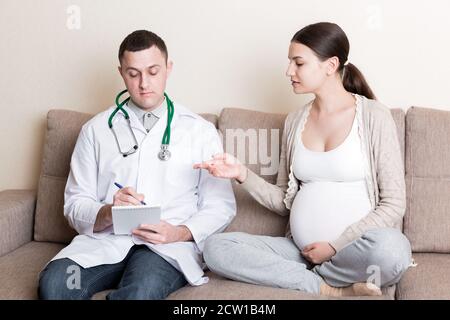 Der männliche Arzt schreibt Beschwerden von schwangeren Frauen in ein Notizbuch. Gesundheitliche Probleme während der Schwangerschaft Konzept. Stockfoto