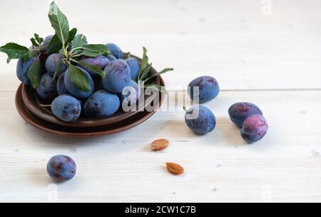 Ein paar reife blaue Pflaumen in einem braunen Teller auf einem weißen Holztisch. Mehrere Knochen liegen getrennt Stockfoto