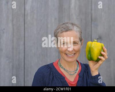 Moderne kaukasische kanadische Countryfrau mittleren Alters mit kurzen Haaren hält eine große organische Paprika in ihrer linken Hand. Stockfoto