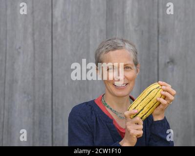 Moderne kaukasische kanadische Countryfrau mittleren Alters mit kurzen Haaren hält einen frischen Bio-Süßkartoffelkürbis in den Händen. Stockfoto