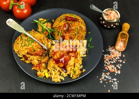 Rotes Risotto mit Tomaten und Chili auf schwarzem Teller. Traditionelle Mahlzeit. Hausmannskost. Würziges Essen. Huhn und Schweinefleisch im Risotto. Stockfoto