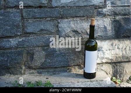 Eine leere Weinflasche aus dunklem Glas mit weißem Etikett und einem Kuhstopper, der auf der Straße gegen eine Steinmauer steht. Foto mit viel Platz für Kopien. Stockfoto