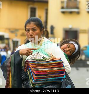 Quito, Ecuador - 2. Dezember 2008: Historische Innenstadt. Nahaufnahme der jungen Mutter mit unruhigen Kind auf dem Rücken, ambulante Verkäufer von gefalteten bunten Text Stockfoto