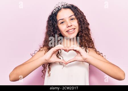 Schönes Kind Mädchen mit lockigen Haaren trägt Prinzessin Tiara lächelnd in der Liebe tun Herz Symbol Form mit Händen. Romantisches Konzept. Stockfoto