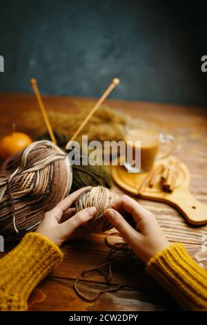 Hände in orange Pullover mit Garn, Stricknadeln und Kaffee auf Holztisch. Handwerk, Hobby. Herbstkomposition für Postkarten und Flyer. Kaffee und Zimtstangen. Betonwand im Hintergrund. Stockfoto