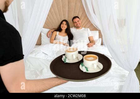 Zimmerservice bringt 2 Tassen Kaffee ins Hotel Zimmer für ein Ehepaar, das in einem Bett im liegt Morgen Stockfoto