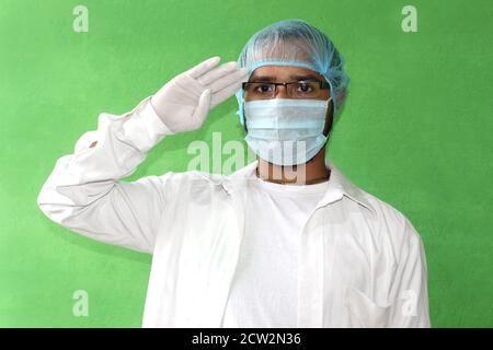 Ein junger Arzt in medizinischer Uniform mit einer schützenden Gesichtsmaske und Handschuhen salutieren während covid-19, Corona-Virus. Stockfoto