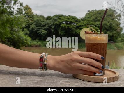 Weibliche Hand hält Eishonig Zitrone Kaffee. Ein Glas Kaffee gemischt mit Honig und Zitrone mit Blick auf den Fluss. Erfrischendes Sommergetränk-Konzept. Selektiv Stockfoto