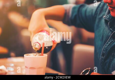 Frau Hand halten pour Cola in Glas Cola spritzt Mit Eis auf Holztisch Cafe Hintergrund Stockfoto