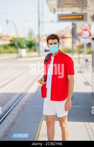 Junger Mann trägt eine chirurgische Maske während der Wartezeit auf ein Zug an einem Außenbahnhof Stockfoto