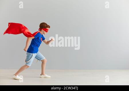 Kleiner Junge in einem Superhelden-Kostüm, der auf einem Hintergrund einer grauen Wand läuft. Stockfoto