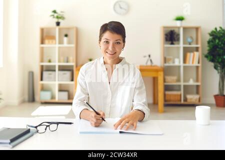 Lächelnde junge Frau, die sich Notizen gemacht hat, während sie einem Online-Vortrag in einem hellen Raum zuhört. Stockfoto