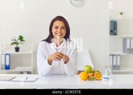 Glückliche weibliche Ernährungsberaterin, die im Büro an ihrem Schreibtisch saß und schaute Vor der Kamera und lächelnd Stockfoto