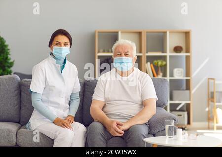 Portrait des Therapeuten und des gealterten weißhaarigen Mannes, beide mit medizinischen Gesichtsmasken, sitzend auf der Couch zu Hause. Stockfoto