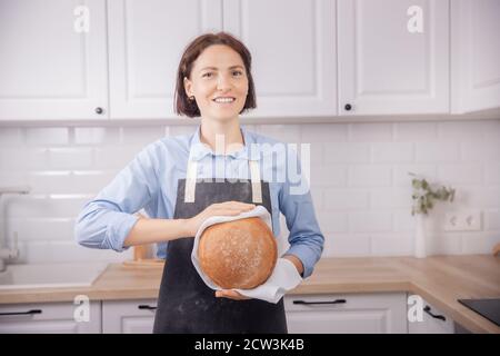 Die Bäckerin in der Schürze hält das runde Brot in den Händen Hintergrund der weißen Küche im skandinavischen Stil Stockfoto